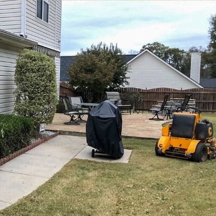Side view of backyard with a grill and yellow riding mower around patio.